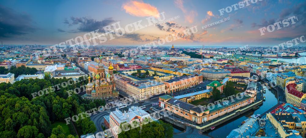 В центре изображения находится знаменитый Спас на Крови с его красочными луковичными куполами, окруженный многочисленными каналами и зеленью городских парков. Мягкий свет и пастельные тона неба создают успокаивающую и вдохновляющую атмосферу. Этот снимок идеален для туристических брошюр, культурных публикаций, учебных материалов по искусствоведению и архитектуре, а также в качестве фотообоев или декоративных панно.